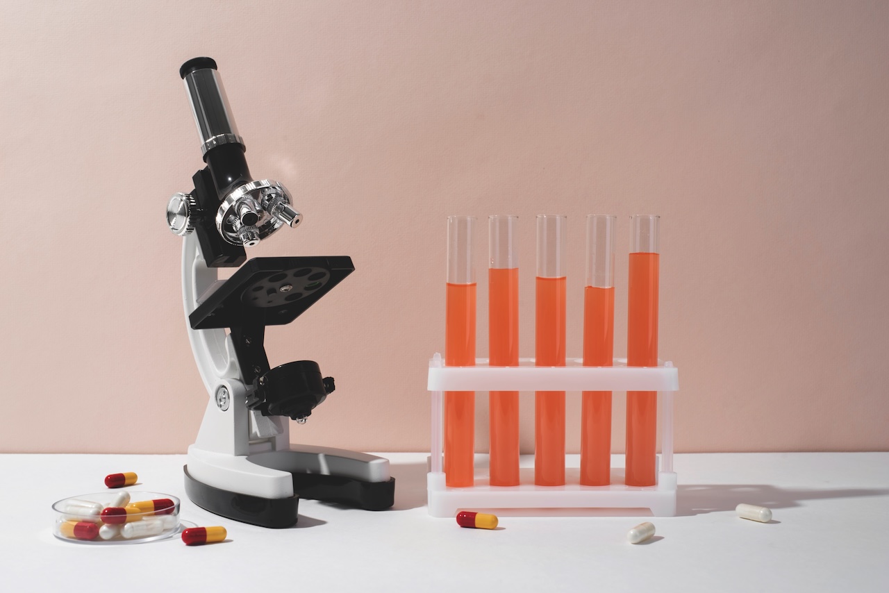 Stock photo of a optical microscope with five test tubes containing an orange solution and some red and yellow pills on the table.