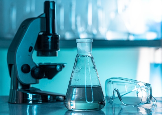 Stock photo of part of a laboratory table with an optical microscope, an Erlenmeyer flask, and safety glasses.